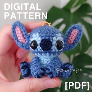 Può includere: Un personaggio blu all'uncinetto fatto a mano con grandi occhi neri, orecchie interne rosa e un portachiavi color oro. Il testo "DIGITAL PATTERN" e "[PDF]" sono visibili nell'immagine. Il personaggio è un popolare personaggio dei cartoni animati.