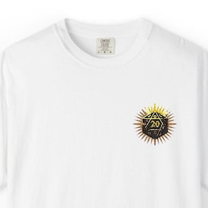Può includere: T-shirt bianco con scollo tondo e grafica dorata dettagliata di un dado a venti facce. Il dado è centrato sul petto. La maglietta ha l'etichetta del marchio "Comfort Colors".