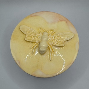 May include: A round, cream-coloured ceramic lid featuring a raised, embossed bee design in the centre. The surface has a glossy, glazed finish with subtle, light brown marbled variations and a smooth, circular edge surrounding the insect motif.