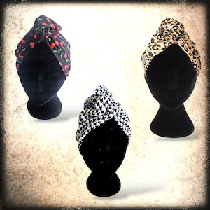May include: Three hair turbans displayed on black mannequins. The turbans are made of fabric with different patterns: red cherries on black, black and white houndstooth, and brown and tan leopard print.