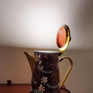 May include: A vintage-style metal teapot with a floral pattern in shades of brown, orange, and white. The teapot has a gold-coloured handle and spout, and the lid is open, revealing a light inside.