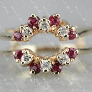 Op de afbeelding: Een goudkleurige ringbeschermer met een halo-ontwerp. De ring heeft afwisselend ronde, rode edelstenen en heldere, ronde edelstenen. De edelstenen zijn gezet in een delicaat, bloemachtig patroon.