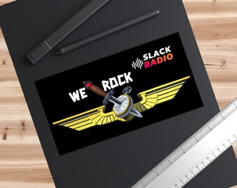 Op de afbeelding: Een rechthoekige sticker met de tekst WE ROCK en SLACK RADIO en een afbeelding van een dolk door een zakhorloge boven gele vleugels. Een zwarte stylus en een liniaal van 0 tot 15 cm liggen in de buurt.