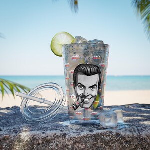 May include: A clear plastic tumbler with a lid and straw, filled with ice and a lime wedge. The tumbler features a black and white illustration of a man with a pipe and the text "Slack Radio". The background shows a beach scene.