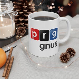 May include: White ceramic mug with the text "prg gnus" in a black font. The letters "prg" are in red, black, and blue squares. The mug is filled with a dark liquid and sits on a light gray surface.