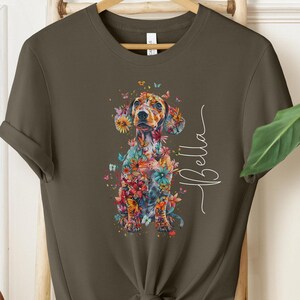 Op de afbeelding: Een groen T-shirt met een kleurrijke illustratie van een teckel met bloemen en vlinders. De tekst "Bella" staat in witte cursieve letters aan de rechterkant van de afbeelding.