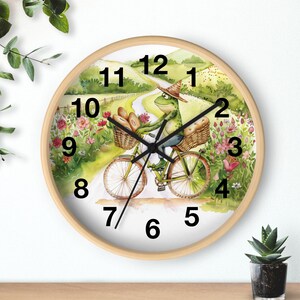 May include: A round wall clock with a white background and a green frog riding a bicycle with baskets of bread. The clock face has black numbers 1 through 12 and a black hand pointing to the number 12.