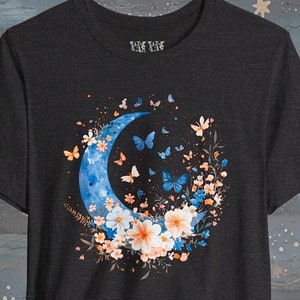Op de afbeelding: Een donkergrijs T-shirt met een blauwe halve maan omringd door roze en witte bloemen en blauwe vlinders.