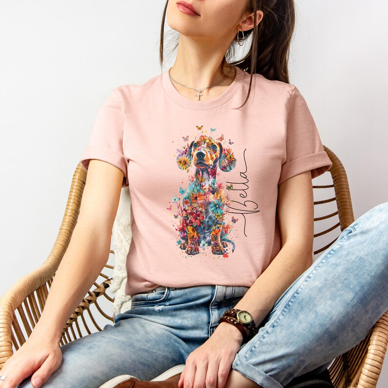 Op de afbeelding: Een roze T-shirt met een bloemenprint van een teckel en de naam "Bella" op de voorkant.