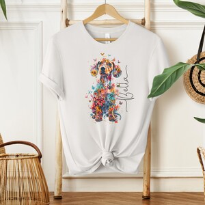 Op de afbeelding: Wit T-shirt met een kleurrijk bloemig teckelontwerp en de naam "Bella" gedrukt op de voorkant.