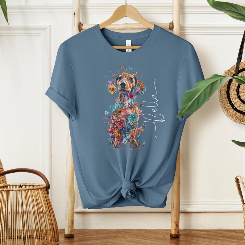 Op de afbeelding: Een blauw t-shirt met een bloemig teckelontwerp en de naam "Bella" in wit gedrukt.