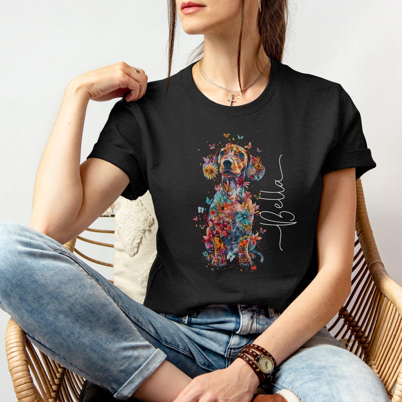 Op de afbeelding: Zwart T-shirt met een kleurrijke, bloemige teckel-illustratie en de naam "Bella" in cursief.