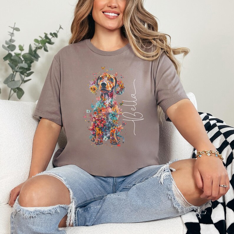 Op de afbeelding: Een grijs T-shirt met een bloemen-dachshund ontwerp en de naam "Bella" gedrukt op de rechterkant van het shirt.