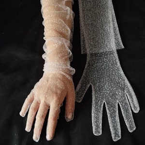May include: A pair of white sheer gloves with a sparkly finish. The gloves are long and have a ruffled design. One glove is shown on a hand, while the other is laid flat.