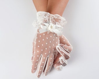 Puede incluir: Guantes de encaje blanco con un pequeño detalle de flor blanca y un lazo. Los guantes tienen un patrón de lunares.