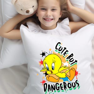 Puede incluir: Almohada blanca con una imagen de dibujos animados de Piol&iacute;n, un canario amarillo, con el texto "Cute But Dangerous" en negro y verde. La almohada est&aacute; sobre una cama blanca.