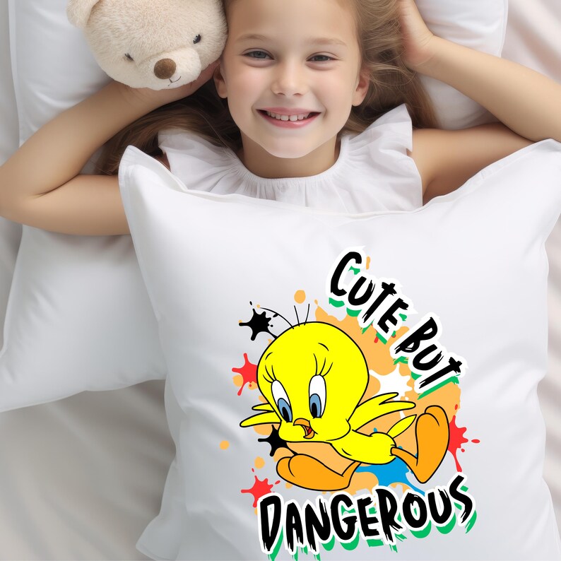 Puede incluir: Almohada blanca con una imagen de dibujos animados de Piol&iacute;n, un canario amarillo, con el texto "Cute But Dangerous" en negro y verde. La almohada est&aacute; sobre una cama blanca.