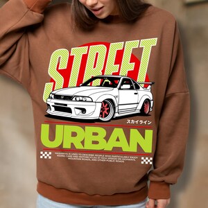 Puede incluir: Sudadera marr&oacute;n con un gr&aacute;fico de un coche deportivo blanco con ruedas rojas, sobre un c&iacute;rculo rojo y las palabras "STREET" y "URBAN" en verde. Tambi&eacute;n est&aacute; presente el texto "HASHIRIYA IS USED TO DESCRIBE PEOPLE WHO PARTICULARLY ENJOY RIDING CARS AND MOTORCYCLES AT HIGH SPEEDS ON HIGHWAYS, MOUNTAIN ROADS, AND OTHER PUBLIC ROADS".