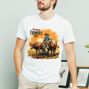 Pu&ograve; includere: T-shirt bianca con un'illustrazione colorata per la Festa del Contadino. La grafica raffigura un contadino con due buoi che ara un campo al tramonto. Il testo "Happy Farmer's Day" &egrave; visualizzato sopra.