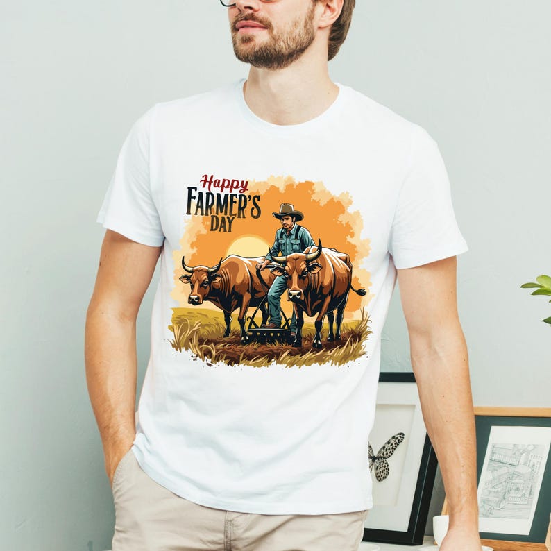 Pu&ograve; includere: T-shirt bianca con un'illustrazione colorata per la Festa del Contadino. La grafica raffigura un contadino con due buoi che ara un campo al tramonto. Il testo "Happy Farmer's Day" &egrave; visualizzato sopra.