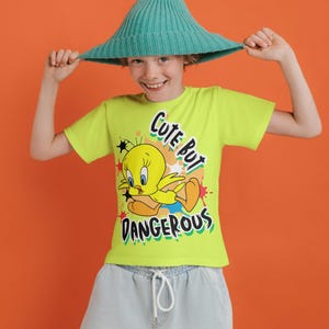 Puede incluir: Una camiseta verde lima con Piol&iacute;n y el texto "Cute But Dangerous". El personaje de dibujos animados es amarillo con pies naranjas y est&aacute; sobre estrellas rojas. La persona lleva un sombrero turquesa.