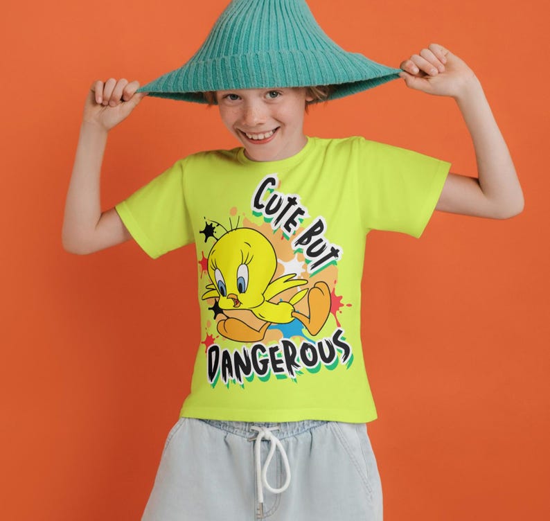 Puede incluir: Una camiseta verde lima con Piol&iacute;n y el texto "Cute But Dangerous". El personaje de dibujos animados es amarillo con pies naranjas y est&aacute; sobre estrellas rojas. La persona lleva un sombrero turquesa.