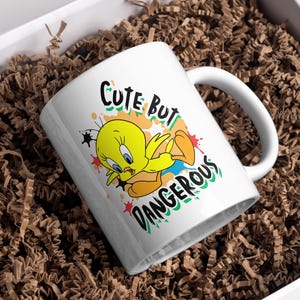 Puede incluir: Taza de cer&aacute;mica blanca con Tweety Bird y el texto "CUTE BUT DANGEROUS". El personaje de dibujos animados es amarillo con ojos azules y pies naranjas. La taza est&aacute; dentro de una caja blanca llena de material de embalaje marr&oacute;n.