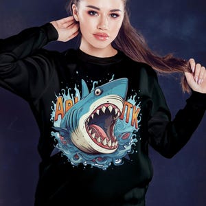 May include: Black sweatshirt with a graphic print of a blue shark with its mouth open, surrounded by water. The text "Акула" is printed on the sweatshirt.