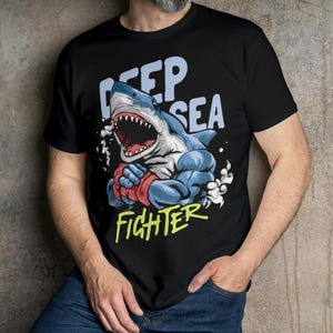 May include: Black t-shirt featuring a cartoon illustration of a muscular shark wearing boxing gloves. The text "Deep Sea Fighter" is printed on the shirt.