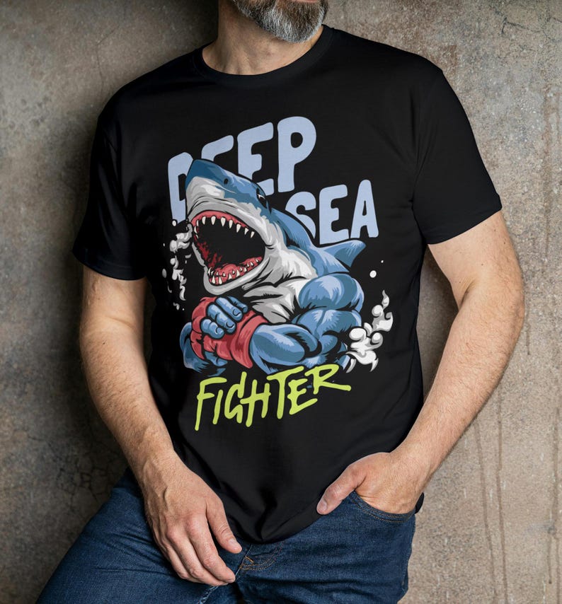 May include: Black t-shirt featuring a cartoon illustration of a muscular shark wearing boxing gloves. The text "Deep Sea Fighter" is printed on the shirt.