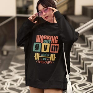 May include: Black sweatshirt with the text "WORKING OUT GYM MY THERAPY" in a colorful design. The graphic includes a barbell and the text "8 lbs 6 lbs". The sweatshirt is worn by a person wearing pink glasses.