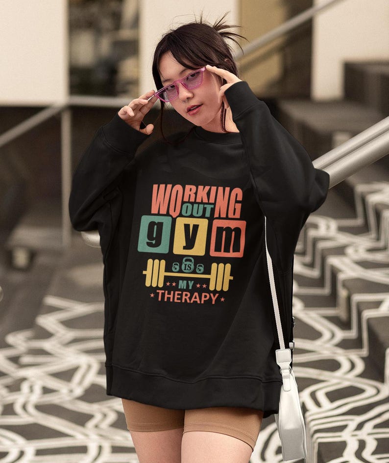 May include: Black sweatshirt with the text "WORKING OUT GYM MY THERAPY" in a colorful design. The graphic includes a barbell and the text "8 lbs 6 lbs". The sweatshirt is worn by a person wearing pink glasses.