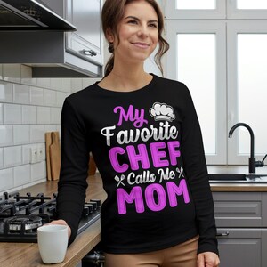 May include: Black long-sleeve shirt with the text "My Favorite CHEF Calls Me MOM" in white and pink. The shirt features a chef's hat graphic. The woman is holding a white mug.