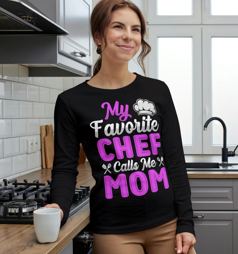 May include: Black long-sleeve shirt with the text "My Favorite CHEF Calls Me MOM" in white and pink. The shirt features a chef's hat graphic. The woman is holding a white mug.