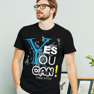 May include: Black t-shirt with a graphic design that says "Yes You Can!" in white, blue, and yellow lettering with gray paint splatters. The text "Free Style" is printed below the graphic.