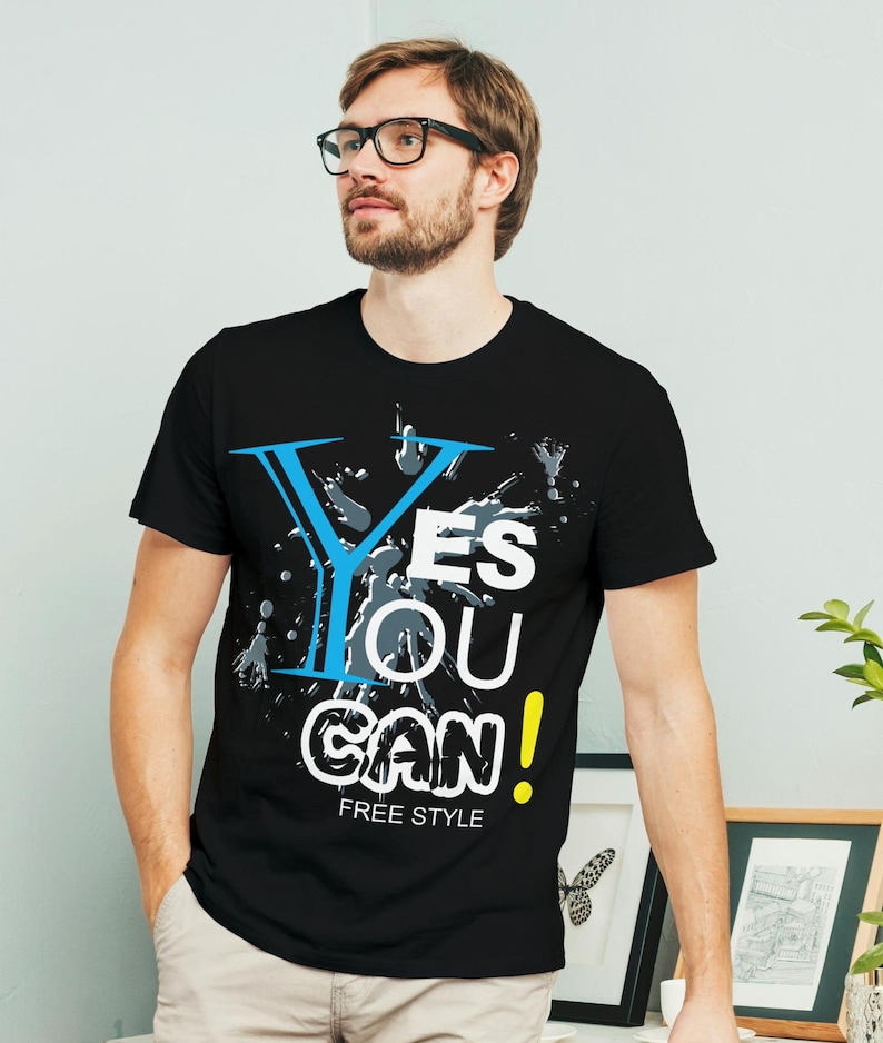 May include: Black t-shirt with a graphic design that says "Yes You Can!" in white, blue, and yellow lettering with gray paint splatters. The text "Free Style" is printed below the graphic.