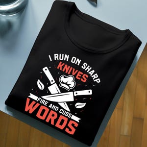 May include: Black t-shirt with a chef-themed graphic. The design includes knives, a chef's hat, and the text "I RUN ON SHARP KNIVES FIRE AND CUSS WORDS" in white and red. The shirt is folded neatly on a light blue surface.