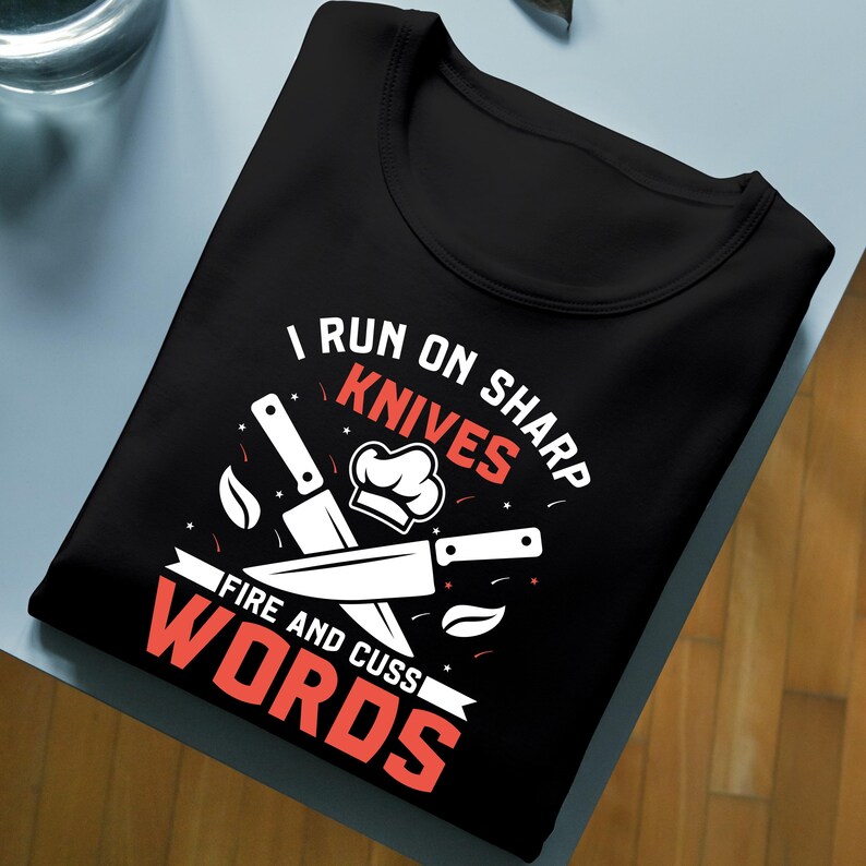 May include: Black t-shirt with a chef-themed graphic. The design includes knives, a chef's hat, and the text "I RUN ON SHARP KNIVES FIRE AND CUSS WORDS" in white and red. The shirt is folded neatly on a light blue surface.