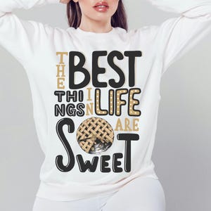 May include: A white sweatshirt featuring the phrase "THE BEST THINGS IN LIFE ARE SWEET" in black, gold, and white. The design includes a pie graphic. This is a casual, comfortable style of clothing.
