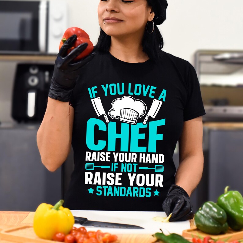 May include: Black t-shirt with the text "IF YOU LOVE A CHEF RAISE YOUR HAND IF NOT RAISE YOUR STANDARDS" in white and teal. The shirt features a chef's hat and cleavers. A red bell pepper is held in the left hand.