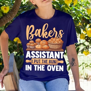 May include: A navy blue t-shirt featuring the text "Bakers Assistant" and "I put the bun in the oven". The design includes illustrations of baked goods, such as muffins, bread, and a basket of rolls, along with a rolling pin graphic.
