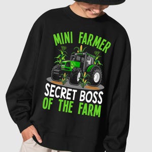 Pu&ograve; includere: Felpa nera con grafica di un trattore verde e la scritta "MINI FARMER SECRET BOSS OF THE FARM". Il design include piante di mais e un trattore verde.