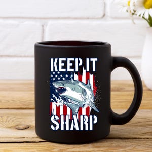 May include: Black ceramic coffee mug featuring a white shark graphic and the text "KEEP IT SHARP" in white lettering. The shark is swimming in front of an American flag.