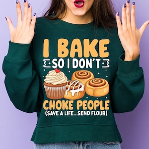 May include: A dark green sweatshirt featuring the text "I BAKE SO I DON'T CHOKE PEOPLE (SAVE A LIFE...SEND FLOUR)". The design includes a cupcake and cinnamon rolls in yellow, white, and brown tones.