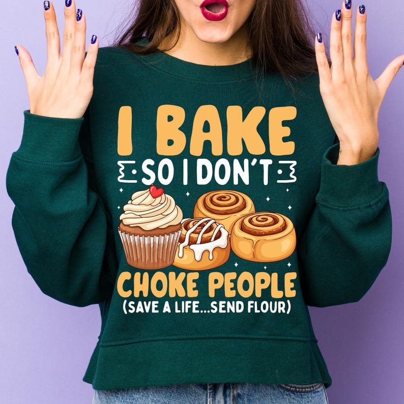 May include: A dark green sweatshirt featuring the text "I BAKE SO I DON'T CHOKE PEOPLE (SAVE A LIFE...SEND FLOUR)". The design includes a cupcake and cinnamon rolls in yellow, white, and brown tones.