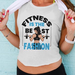 May include: White tank top with the text "FITNESS IS THE BEST FASHION" in black and blue. The graphic features a woman lifting weights. The woman is wearing a black sports bra. The person is wearing black athletic pants.