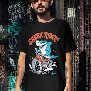 May include: Black t-shirt featuring a graphic of a shark riding a motorbike. The shark is blue and has a toothy grin. The motorbike is red and has a California number plate. The text "SHARK RIDER!" is printed in red on the shirt.