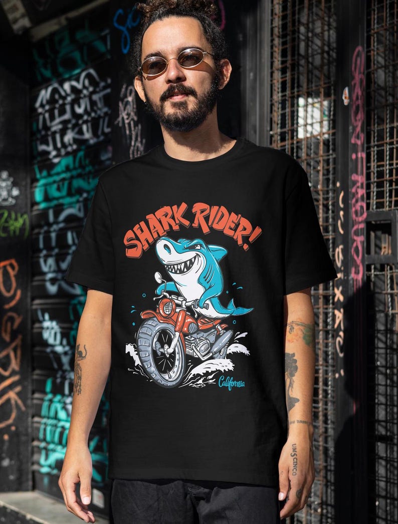 May include: Black t-shirt featuring a graphic of a shark riding a motorbike. The shark is blue and has a toothy grin. The motorbike is red and has a California number plate. The text "SHARK RIDER!" is printed in red on the shirt.
