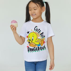 Puede incluir: Camiseta blanca con un dibujo animado de Tweety Bird y el texto "Cute But Dangerous". La camiseta se combina con unos vaqueros azules. Una magdalena con glaseado rosa se sostiene en la mano.