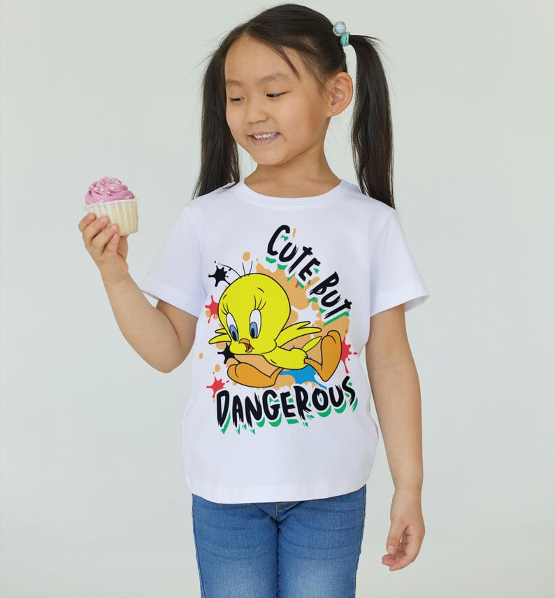 Puede incluir: Camiseta blanca con un dibujo animado de Tweety Bird y el texto "Cute But Dangerous". La camiseta se combina con unos vaqueros azules. Una magdalena con glaseado rosa se sostiene en la mano.